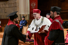 Ceremonia wręczenia dyplomów. Absolwent w czarnej todze z czerwonym kołnierzem i birecie podaje rękę rektorowi w białej todze z łańcuchem i czerwonym biretem. Rektor trzyma czerwoną teczkę. W tle osoby w togach. Na ścianie herb i napis „Warszawski Uniwersytet Medyczny”.