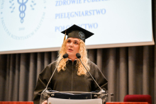 Młoda kobieta w czarnej todze i birecie z emblematem uczelni stoi za mównicą. Ma długie, kręcone blond włosy. W tle ekran z napisem „ZDROWIE PUBLICZNE, PIELĘGNIARSTWO, POŁOŻNICTWO”.