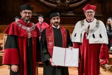 Trzech mężczyzn w togach i biretach stoi na tle drewnianej ściany. Środkowy w czarnej todze z bordową szarfą „Class of 2025 Graduate” trzyma dyplom. Po bokach osoby w czerwonych i czerwono-białych togach z łańcuchami rektorskimi. W tle inni uczestnicy uroczystości.