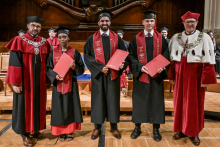 Pięć osób w togach i biretach stoi na scenie. Trzy osoby w środku trzymają czerwone dyplomy. Po bokach osoby w czerwonych togach z ozdobnymi łańcuchami. W tle drewniana dekoracja i siedzący uczestnicy ceremonii.