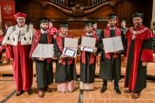 Grupa absolwentów w czarnych togach i biretach pozuje z dyplomami. Po bokach stoją osoby w czerwonych togach z insygniami rektorskimi. W tle drewniany wystrój sali i organy.