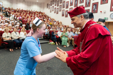 Uroczystość czepkowania absolwentów pielęgniarstwa i położnictwa. Kobieta w niebieskim uniformie medycznym i czepku położnych stoi na scenie i ściska dłoń starszego mężczyzny w czerwonym stroju akademickim. 