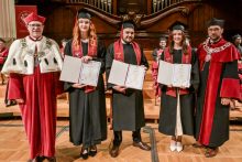 Pięć osób w togach i biretach stoi na drewnianej podłodze. Trzy osoby w środku mają czarne togi z czerwonymi szarfami i trzymają dyplomy. Po bokach osoby w czerwonych togach z białymi elementami i łańcuchami. W tle drewniany organ i czerwona tablica z herbem uczelni.
