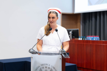 Uroczystość czepkowania absolwentów pielęgniarstwa i położnictwa. Kobieta w białym uniformie medycznym i czepku położnych stoi za mównicą z dwoma mikrofonami.