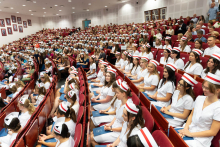 Uroczystość czepkowania absolwentów pielęgniarstwa i położnictwa. W dużej sali siedzą kobiety w uniformach medycznych i czepkach pielęgniarek i położnych, trzymające niebieskie teczki. 