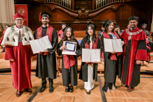 Sześć osób w togach i biretach stoi na scenie. Cztery osoby w środku trzymają dyplomy. Po bokach osoby w czerwonych togach z białymi elementami i łańcuchami rektorskimi. W tle drewniana ściana z organami. Drewniana podłoga.