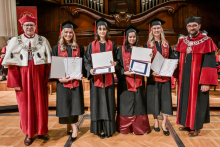 Sześć osób w togach i biretach stoi na drewnianej podłodze. Cztery osoby w środku mają czarne togi z czerwonymi szarfami i trzymają dyplomy. Po bokach osoby w czerwonych togach z białymi elementami i łańcuchami. W tle drewniany organ i czerwona tablica z herbem uczelni.