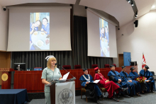 Uroczystość czepkowania absolwentów pielęgniarstwa i położnictwa. Kobieta w zielonym uniformie medycznym i czepku pielęgniarskim stoi za mównicą z dwoma mikrofonami, trzymając kartki papieru. 