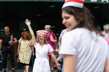 Kobieta w białym uniformie medycznym i czepku położnych stoi na zewnątrz budynku z uniesioną ręką. Na pierwszym planie inna kobieta w białym uniformie medycznym i czepku położnych patrzy w jej kierunku. 