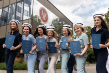 Siedem kobiet w uniformach medycznych i czepkach pielęgniarskich stoi przed budynkiem z dużym logo Uniwersytetu Medycznego w Warszawie. Każda z nich trzyma w rękach granatową teczkę.