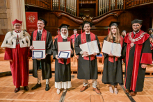 Sześć osób stoi na scenie w auli. Cztery osoby w środku mają czarne togi i birety, trzymają dyplomy. Po bokach osoby w czerwonych togach z insygniami rektorskimi. W tle duży drewniany organ i siedzący uczestnicy ceremonii.