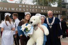 Przed budynkiem Warszawskiego Uniwersytetu Medycznego stoją cztery osoby. Kobieta w uniformie medycznym i czepku pielęgniarskim trzyma granatową teczkę oraz bukiet kwiatów. Obok niej stoi kobieta trzymająca dużego pluszowego misia oraz mężczyzna w granatowej todze akademickiej.