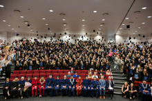 Duża grupa absolwentów w czarnych togach. Nad nimi unoszą się birety rzucone w górę. Na pierwszym planie widać osoby w oficjalnych strojach akademickich. Sala jest pełna.