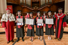 Czworo absolwentów w czarnych togach i biretach stoi na scenie, trzymając dyplomy. Po bokach dwaj mężczyźni w czerwonych togach z insygniami rektorskimi. W tle drewniany prospekt organowy.