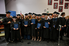 Grupa absolwentów w czarnych togach i biretach stoi w rzędzie, trzymając dyplomy. Wszyscy uśmiechają się do zdjęcia. W tle widoczne są portrety na ścianie i osoby siedzące.