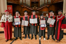 Sześcioro absolwentów w czarnych togach i biretach trzyma dyplomy. Stoją na tle drewnianych organów w sali koncertowej. Po bokach dwaj mężczyźni w czerwonych togach z insygniami rektorskimi.