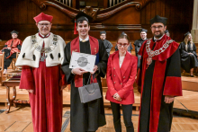 Cztery osoby pozują na scenie. Trzy mają togi i birety, jedna czerwoną marynarkę, a jedna czarną togę z czerwoną szarfą. Osoba w środku trzyma dyplom z napisem „Calvin Radu” i torbę prezentową. W tle drewniane elementy sceny i osoby w togach siedzące na krzesłach.