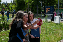 Trzy kobiety stoją na trawie i robią sobie selfie czerwonym telefonem. Dwie z nich trzymają niebieskie teczki. W tle widać zielone drzewa, znak drogowy i tablicę informacyjną.