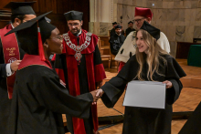 Studentka w czarnej todze i birecie z czerwonym akcentem podaje rękę osobie wręczającej dyplom. Osoba ta ma czarną togę i trzyma biały dokument. W tle widać uczestników ceremonii w togach z czerwonymi elementami i ozdobnymi łańcuchami.