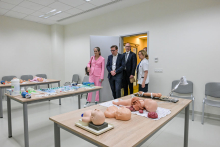 Sala szkoleniowa z dwoma stołami pełnymi fantomów medycznych i narzędzi. Przy drzwiach stoją cztery osoby w formalnych strojach.