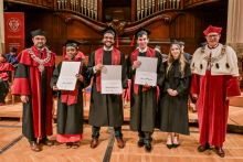 Sześć osób w togach i biretach stoi na scenie. Trzy osoby w środku trzymają dyplomy. Po lewej osoba w czerwonej todze z łańcuchem. Po prawej osoba w czerwonej todze z futrzanym kołnierzem. W tle duże drewniane organy.