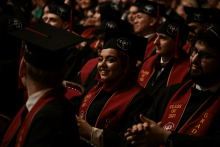 Grupa absolwentów w czarnych togach i biretach siedzi podczas ceremonii ukończenia szkoły. Mają bordowe szarfy z napisem „CLASS OF 2025”. Twarze są zamazane. W tle widoczni są inni uczestnicy wydarzenia.