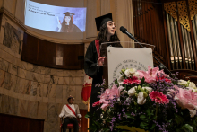 Na pierwszym planie podium z mikrofonem i bukietem kwiatów. Za podium stoi osoba w czarnej todze i birecie, przemawia. W tle ekran z napisem „Agendas 6th Year Class Representative Alexandra Pissa” i zdjęciem osoby w birecie. Po lewej siedzi osoba w historycznym stroju.