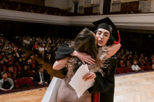 Dwie kobiety obejmują się na scenie podczas ceremonii ukończenia szkoły. Jedna z nich ma czarną togę i czerwony biret, trzyma dyplom. W tle widoczna jest publiczność siedząca w dużej sali.