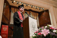 Absolwent w czarnej todze i birecie przemawia na podium z logo Warszawskiego Uniwersytetu Medycznego. W tle widoczne są organy oraz złoto-brązowe zasłony. Po lewej stronie stoi czerwona flaga z herbem uczelni. Na pierwszym planie kwiaty w kolorach różowym, białym i fioletowym.
