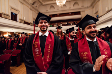 Grupa absolwentów w czarnych togach i biretach stoi w sali koncertowej. Mają czerwone szarfy z napisami „CLASS OF 2025” i „GRADUATE”. W tle widoczny jest balkon z publicznością.