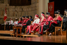 Grupa osób w czerwonych i czarnych togach oraz biretach siedzi na scenie podczas ceremonii akademickiej. W tle marmurowa ściana i sztandar z herbem uczelni. Po lewej stronie osoby ubrane na biało z czerwonymi szarfami.