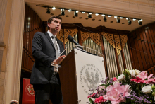 Mężczyzna w garniturze stoi przy mównicy z herbem iWUM. W tle widoczne są organy i dekoracje. Po prawej stronie bukiet kwiatów w kolorach różowym i białym.