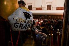 Sala audytoryjna podczas ceremonii ukończenia szkoły. Na pierwszym planie biały balon z napisem „CONGRATS GRAD” i czarną czapką absolwenta. W tle siedzą ludzie, część w togach i biretach. Ściany pokryte drewnianymi panelami, sufit jasny i wysoki.
