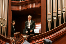 Dwóch mężczyzn w eleganckich garniturach stoi na drewnianym balkonie obok srebrnych piszczałek organowych. Starszy mężczyzna w czarnym smokingu, białej koszuli i muszce stoi na pierwszym planie. Drugi mężczyzna jest częściowo zasłonięty. W tle otwarta książka nutowa na pulpicie. Dominują kolory brązowy i srebrny.