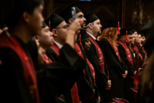 Grupa absolwentów w czarnych togach i biretach stoi w rzędzie. Mają czerwone szarfy z emblematami uczelni. Tło jest ciemne, a światło skierowane jest na absolwentów.
