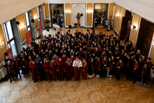 Duża grupa absolwentów w czarnych togach i biretach stoi w eleganckiej sali z drewnianą podłogą i żyrandolami. Trzymają dyplomy i unoszą birety. W tle lustra i osoby obserwujące uroczystość. Na pierwszym planie osoby w czerwonych togach.