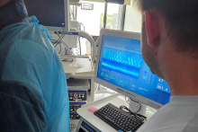 Dwóch pracowników w szpitalnym pomieszczeniu medycznym obserwuje ekran komputera – jeden z nich ma na sobie niebieski fartuch ochronny i rękawiczki, drugi jest ubrany w codzienny str