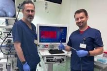 Dwóch pracowników medycznych w niebieskich uniformach stoi obok zaawansowanego sprzętu medycznego. Jeden z nich trzyma cienką rurkę i pokazuje kciuk w górę, drugi stoi przy monitorze. 