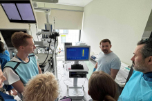 Grupa osób w szpitalnym pomieszczeniu medycznym patrzy na monitor komputera. W tle znajdują się zaawansowane urządzenia diagnostyczne