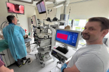 W szpitalnym pomieszczeniu medycznym kilku pracowników wykonuje procedurę przy pacjencie leżącym na łóżku. Jeden z nich, w niebieskim fartuchu ochronnym, trzyma maskę tlenową przy twarzy pacjenta. Inny pracownik w jasnym stroju obserwuje monitor komputera.