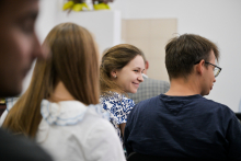 Kilka osób siedzi tyłem w jasnym pomieszczeniu. Na pierwszym planie kobieta w białej bluzce z falbanką. Obok osoba w niebieskiej koszulce, dalej kobieta w białej bluzce z niebieskim wzorem. W tle rozmyte dekoracje i jasna ściana.