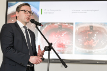 Mężczyzna w garniturze stoi przed mikrofonem i prezentuje slajd na ekranie. 