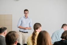 Mężczyzna w okularach występuje przed słuchaczami siedzącymi na krzesłąch