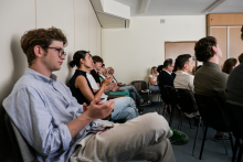 Grupa osób siedzi w sali konferencyjnej, zwrócona twarzami w stronę prelegenta poza kadrem. Ludzie ubrani są nieformalnie. Część osób klaszcze. W tle jasne ściany i drzwi.