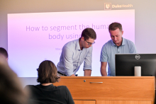 Mężczyzna w okularach oraz mężczyzna prowadzący wykład rozmawiają przy pulpicie. W tle prezentacja na ekranie