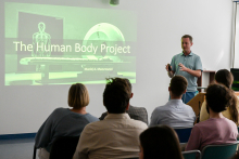 Mężczyzna w niebieskiej koszuli prezentuje projekt „The Human Body Project”. Na ekranie widoczny jest biały szkielet i urządzenie medyczne. W sali jest siedem osób. Pomieszczenie jasne, ze ścianami w kolorze białym.