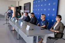 Dwie kobiety i czterech mężczyzn siedzi przy długim stole w sali konferencyjnej. Na stole znajdują się tabliczki z imionami, mikrofony oraz butelki wody. Jeden mężczyzna stoi przy mównicy z mikrofonem i przemawia do publiczności. 