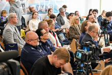 Grupa ludzi w różnych wieku, słuchają wystąpienia. Ludzie siedzą na drewnianych krzesłach. Patrzą przed siebie.