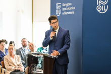 Elegancko ubrany mężczyzna, w okularach i z ciemnymi włosami - prezes naczelnej Rady lekarskiej Łukasz Jankowski. Stoi za mównicą. W prawej ręce trzyma mikrofon i przemawia. W tle niebieska ścianka z białym napisem i logo Okręgowej Izby Lekarskiej. Obok widać kilka osób, które siedzą na krzesłach.