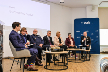 Grupa osób siedzących przy stolikach. Osoby są elegancko ubrane. Rozmawiają. W tle widać ekran multimedialny. Po prawej stronie stoi niebieska ścianka z białymi napisami i z logotypem Okręgowej Izby Lekarskiej.